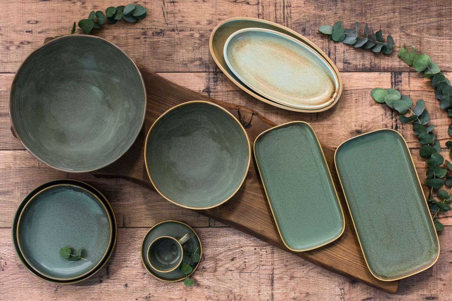 Green ceramic dinnerware set with bowls, plates, and trays on rustic wood surface with eucalyptus leaves.