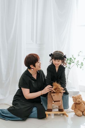 A woman and young girl in matching black outfits smile together beside a rocking horse toy in a bright white room.