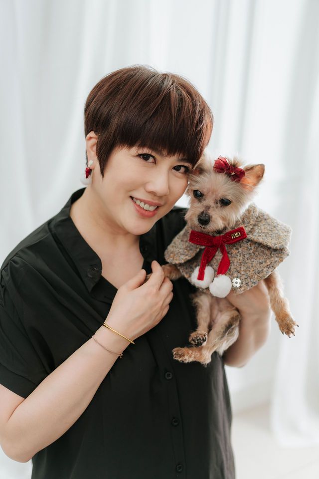 A smiling woman with short hair in a black dress holds a small dog dressed in a festive outfit with a red bow.