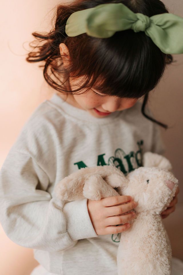 Young girl with green bow headband hugging a plush bunny toy, wearing a gray sweatshirt.