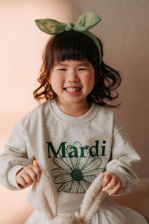 A cheerful young girl wearing a gray Mardi sweatshirt and green bow headband, smiling brightly at the camera.