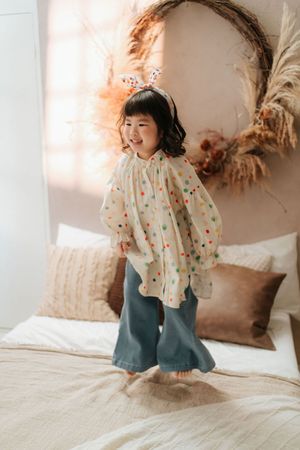 A joyful young girl in a colorful polka dot blouse and teal flare pants jumping on a bed with boho decor.