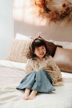 A cheerful young Asian girl sits barefoot on a bed wearing a polka dot blouse, denim skirt, and bunny ear headband.