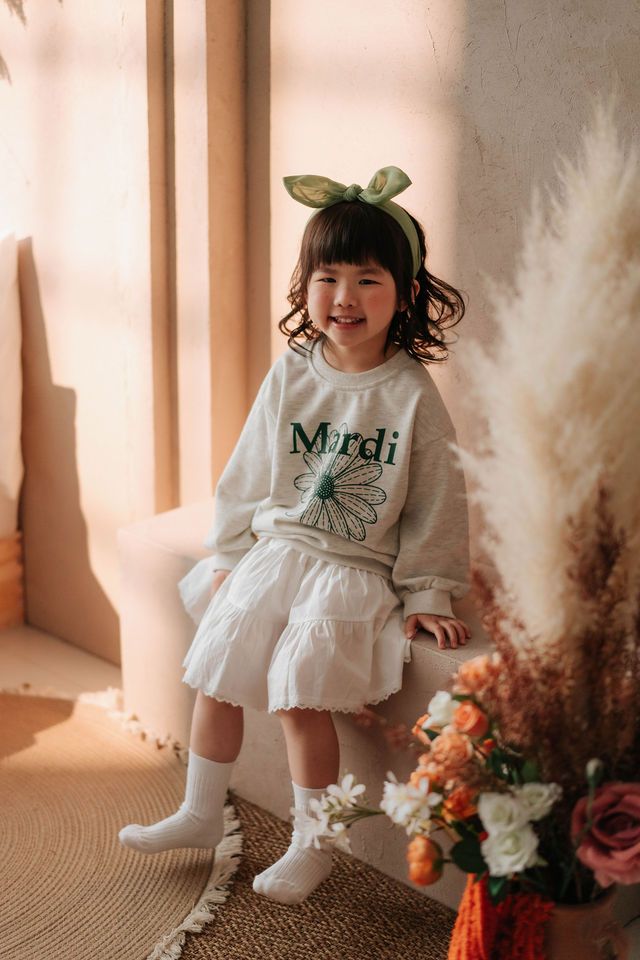 Smiling young girl in gray Mardi sweatshirt, white skirt, and green bow headband, posing near flowers.