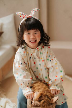 Smiling young Asian girl wearing colorful polka dot blouse and bunny ear headband, holding a fluffy brown animal toy.