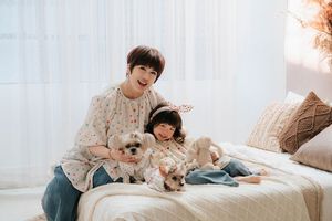Woman and young girl sitting on bed with two small dogs in a bright, cozy room.