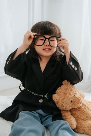 Young girl in oversized black blazer adjusting large glasses beside a teddy bear.