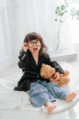 Cute little girl in black blazer laughing while holding glasses next to a teddy bear.