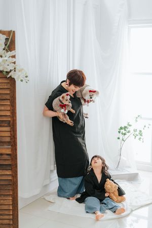 A person in black dress holds two small dogs while a child sits on floor with teddy bear near white curtains.