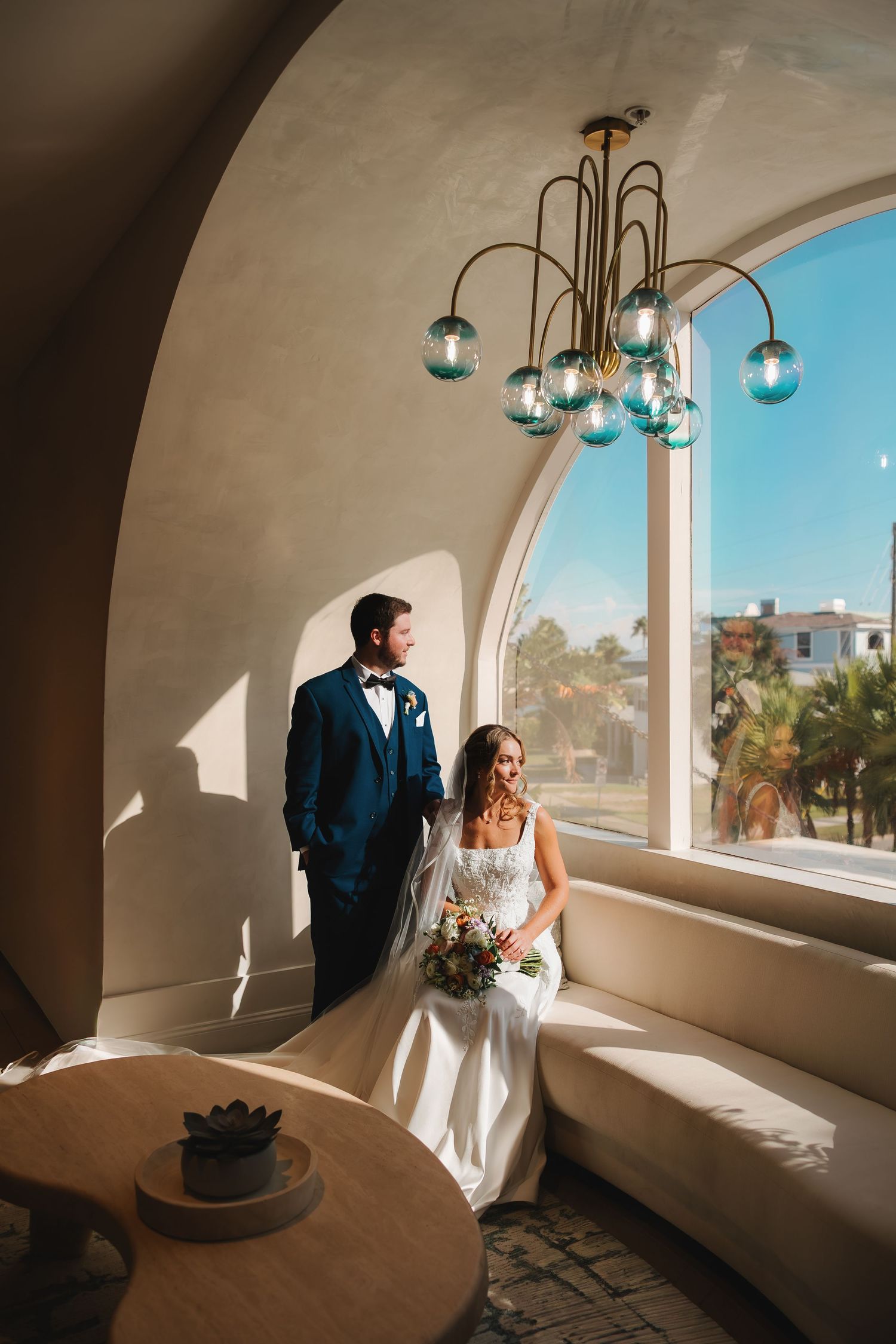 Bride and groom pose in elegant arched alcove with teal chandelier and sunny window backdrop.