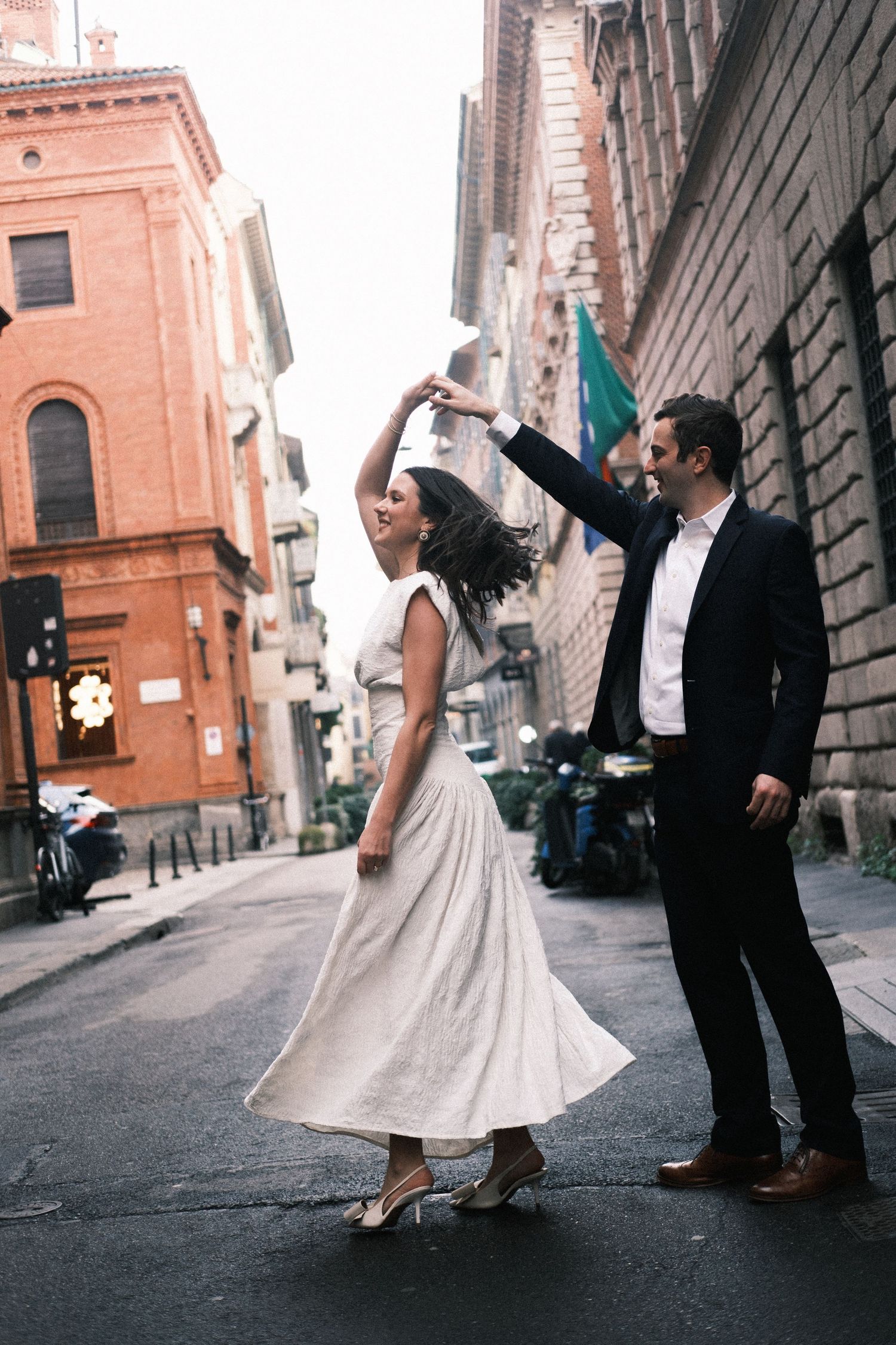 Couple dancing joyfully on a cobblestone street in Italy, woman in white dress being twirled by man in dark suit.