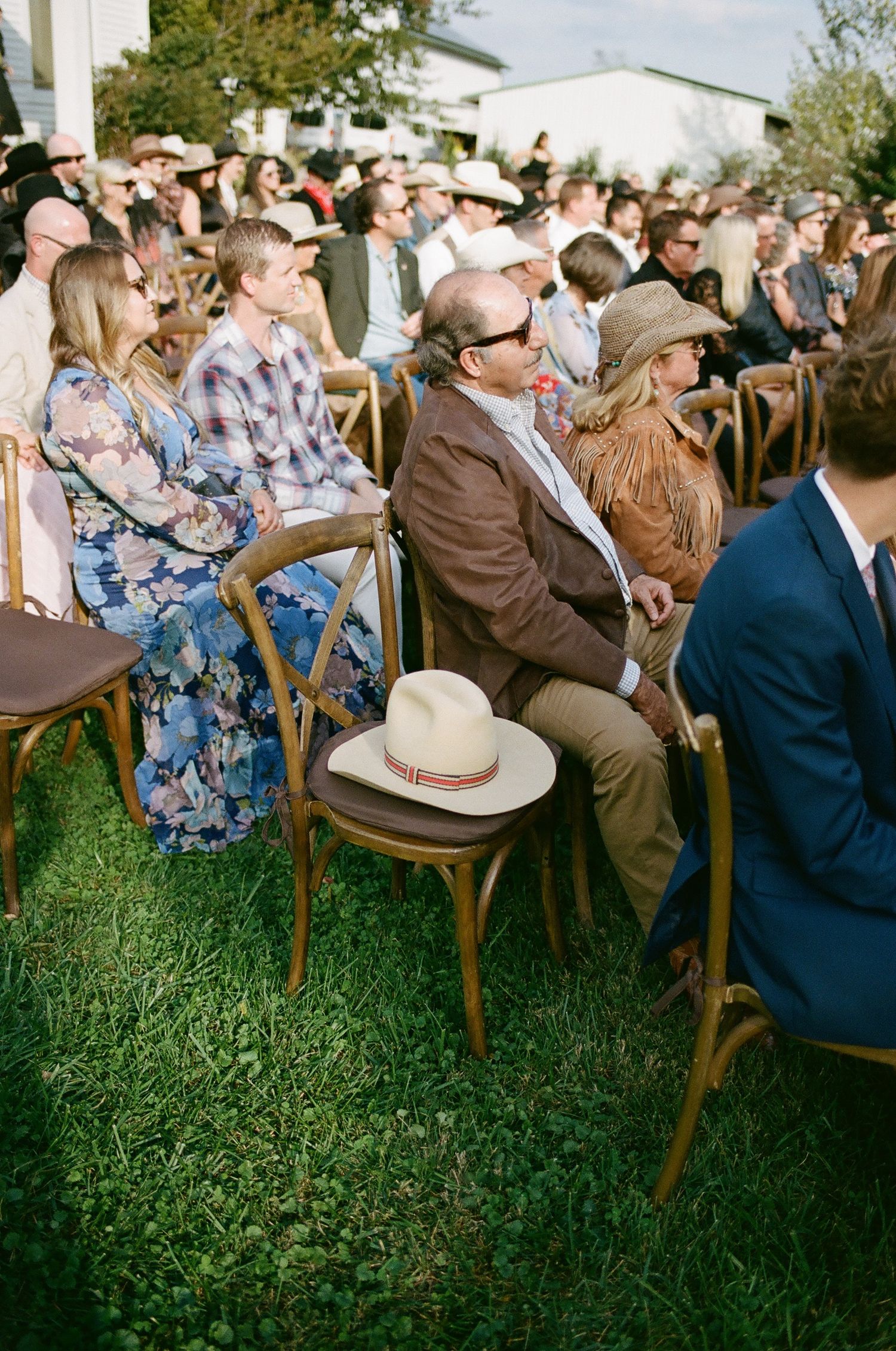 Outdoor wedding guests seated on wooden chairs on grass, man in brown jacket with white cowboy hat on chair beside him.
