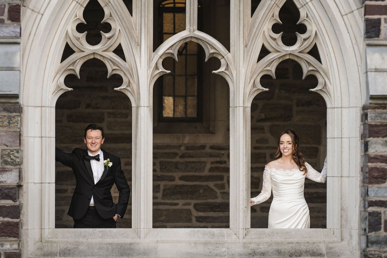 Bride and groom posing in separate Gothic stone arched windows of historic church exterior.