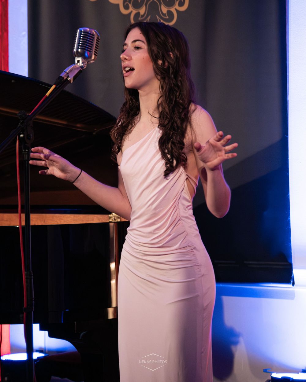 Young woman in elegant white gown singing into vintage microphone on stage at formal event.