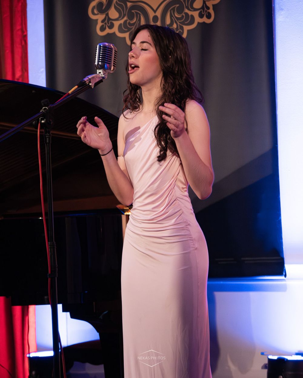 Young woman in pink gown singing into vintage microphone on stage at elegant indoor event.