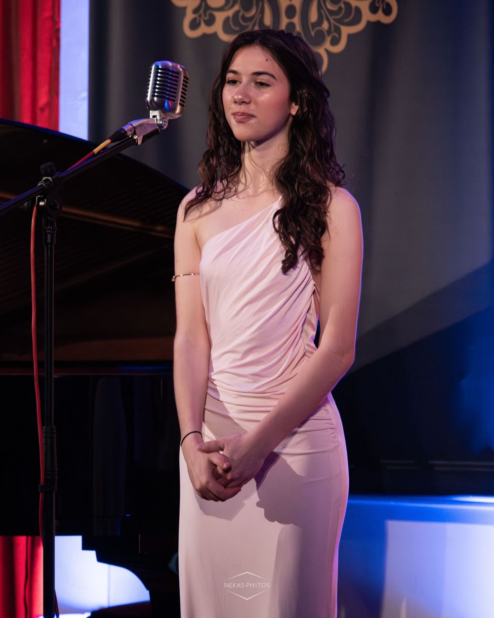 Young woman in elegant pink one-shoulder gown stands at vintage microphone on stage during formal performance event.