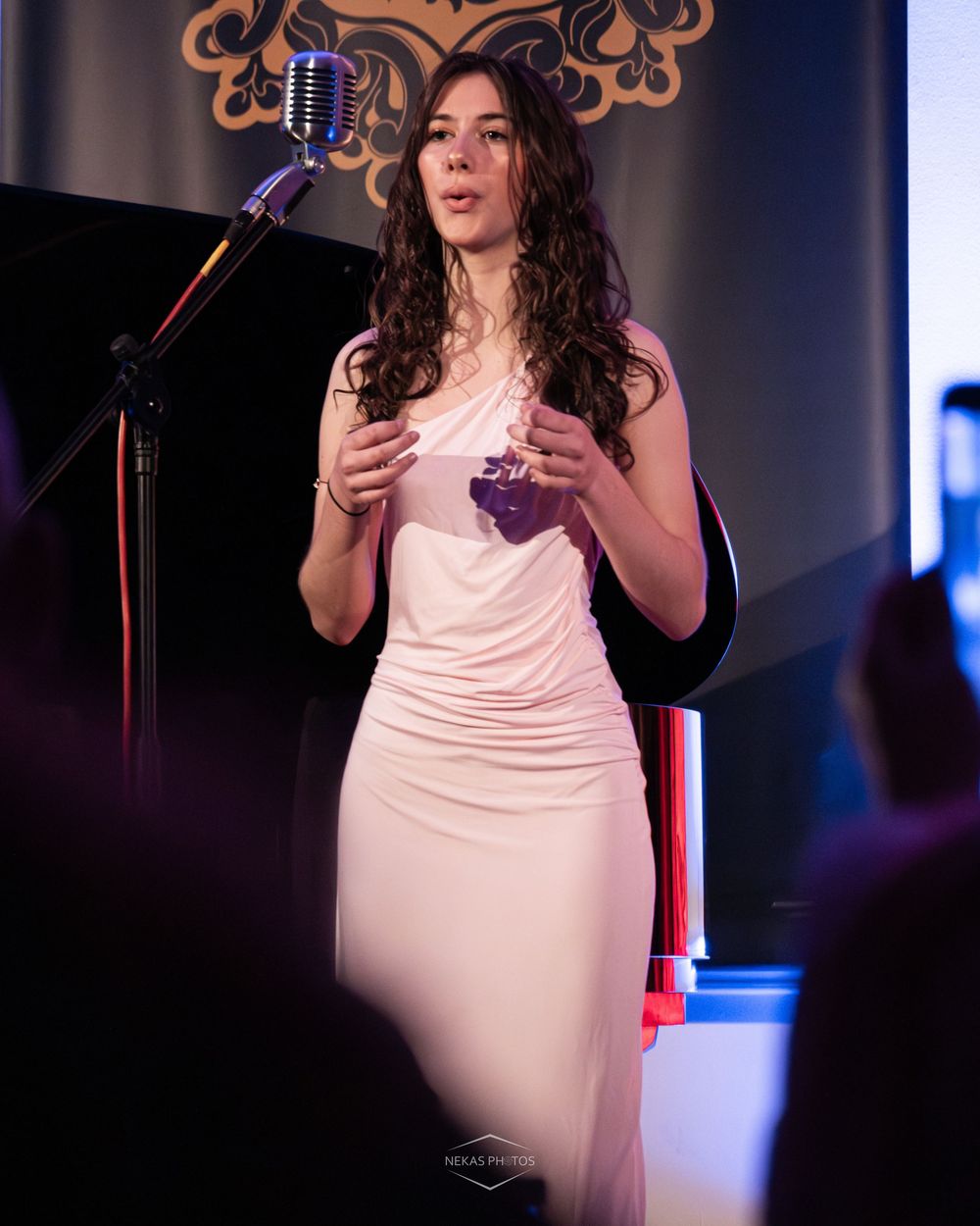 Woman in white dress speaking at microphone stand on stage at formal event.