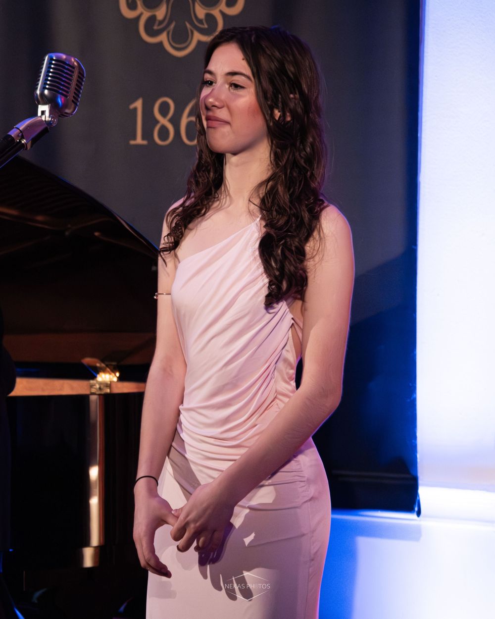Young woman in elegant pink dress stands near vintage microphone at formal event.