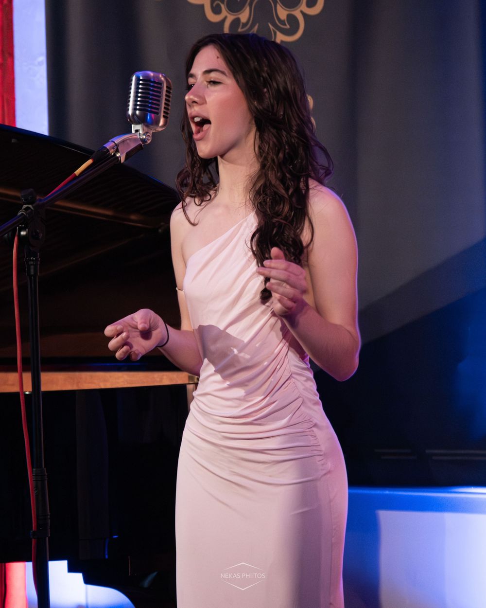Young woman in white dress singing passionately into a vintage microphone on stage at an elegant venue.