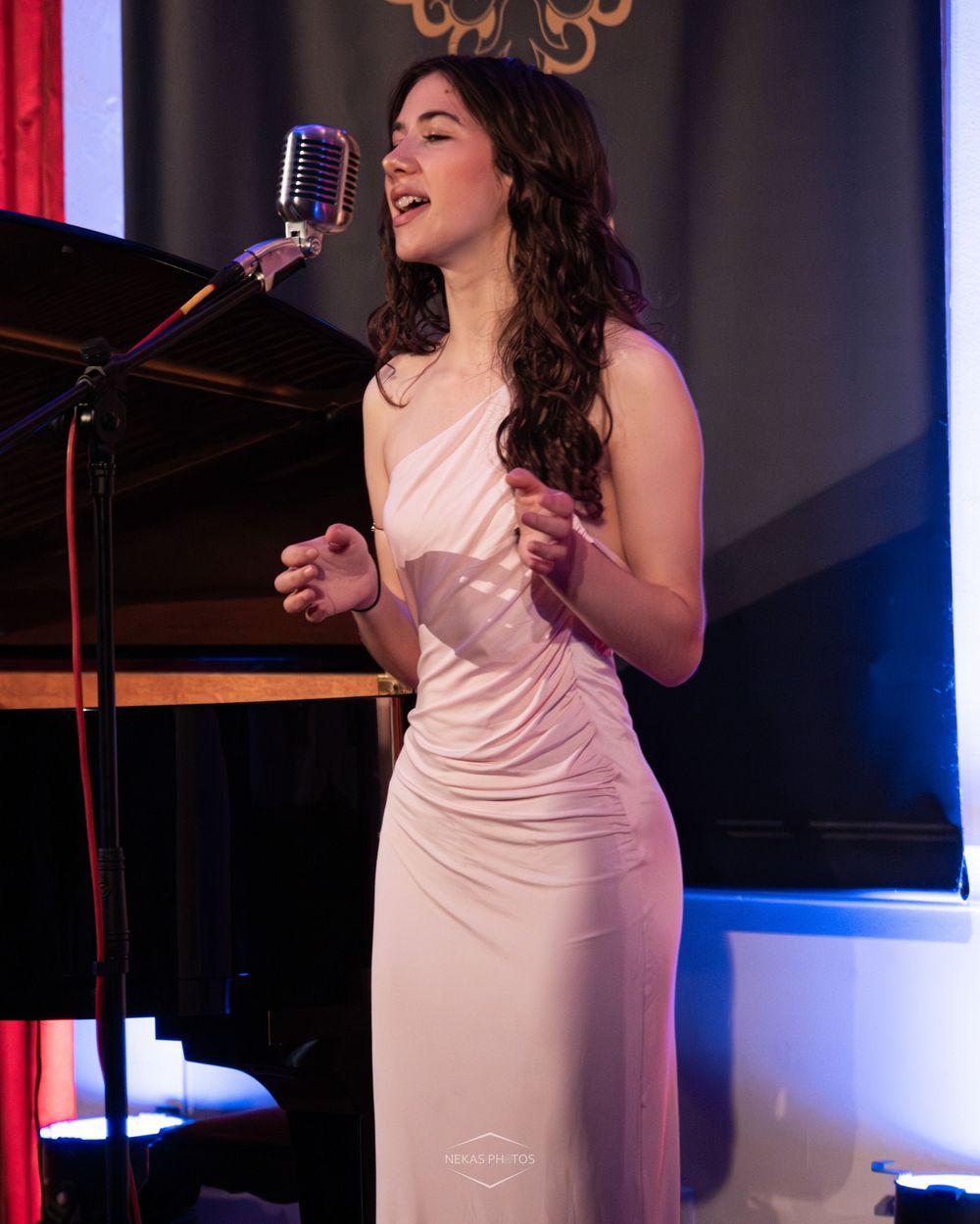 Brunette performer in white dress singing passionately at vintage microphone during live show.
