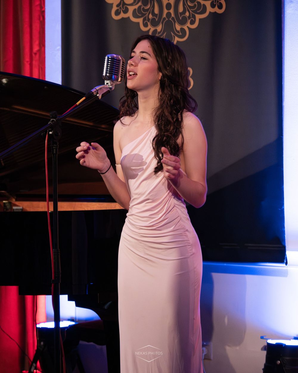 A young woman in a white satin gown sings into a vintage microphone on stage beside a grand piano.