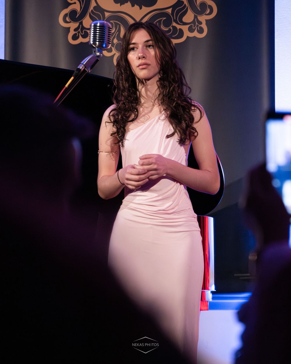 Woman in white dress performing on stage near vintage microphone with curly hair under warm stage lighting.