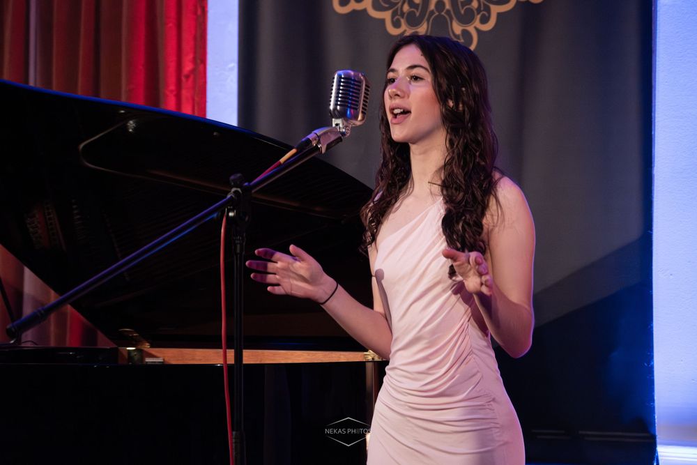 Young woman in white dress singing into vintage microphone on stage beside a grand piano.