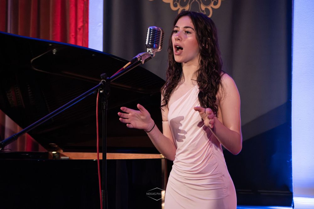 Young woman in white dress singing at vintage microphone on stage during a live performance.
