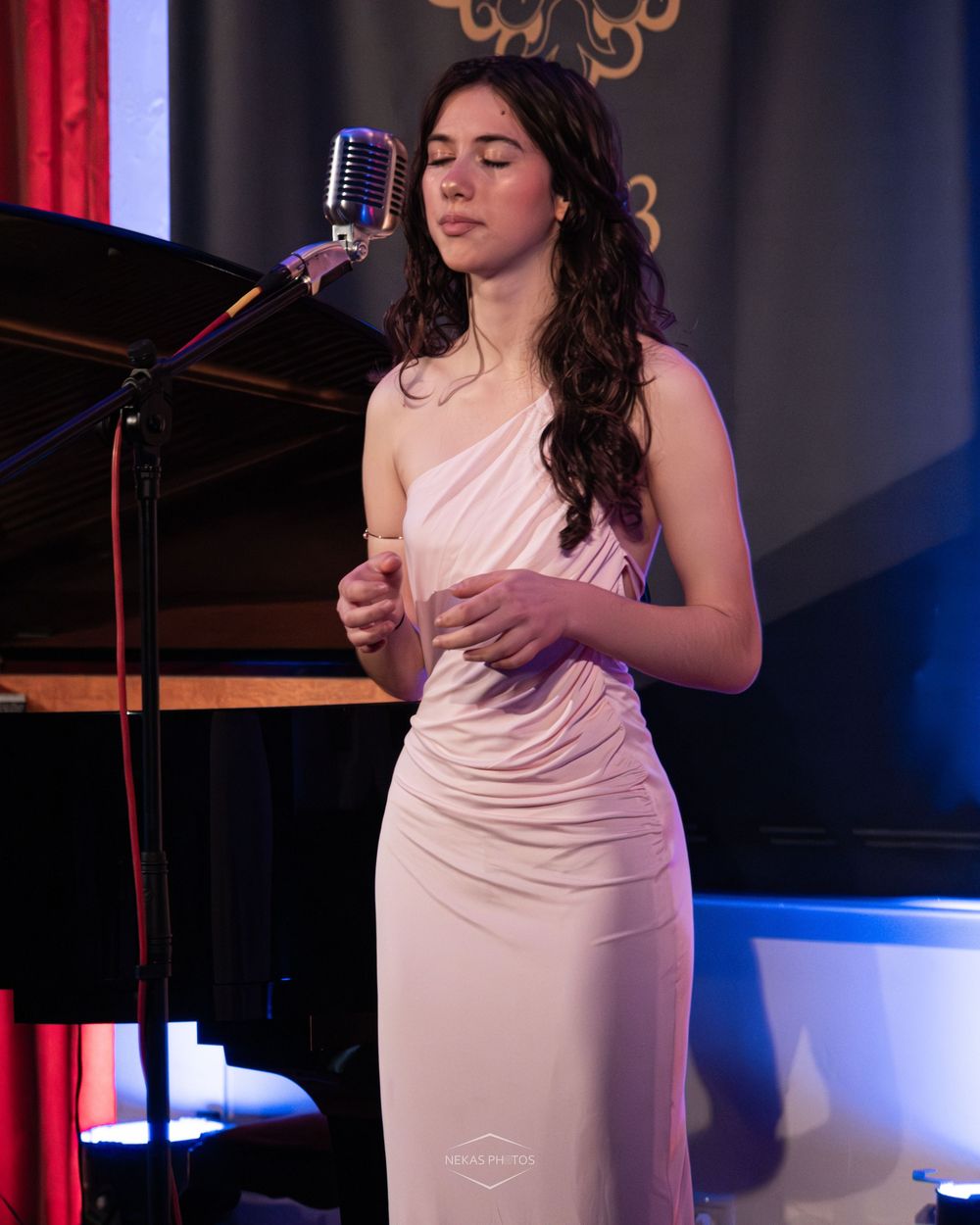 A young woman in a white one-shoulder gown sings at a vintage microphone on an elegant indoor stage.