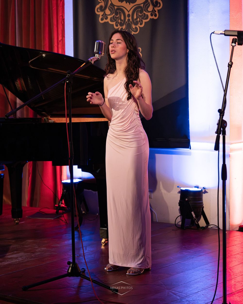 Young woman in elegant white gown singing at vintage microphone on stage with grand piano in background.