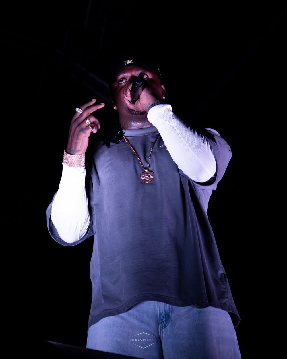 Male hip-hop artist performing live on dark stage wearing grey sweatshirt and gold chain.