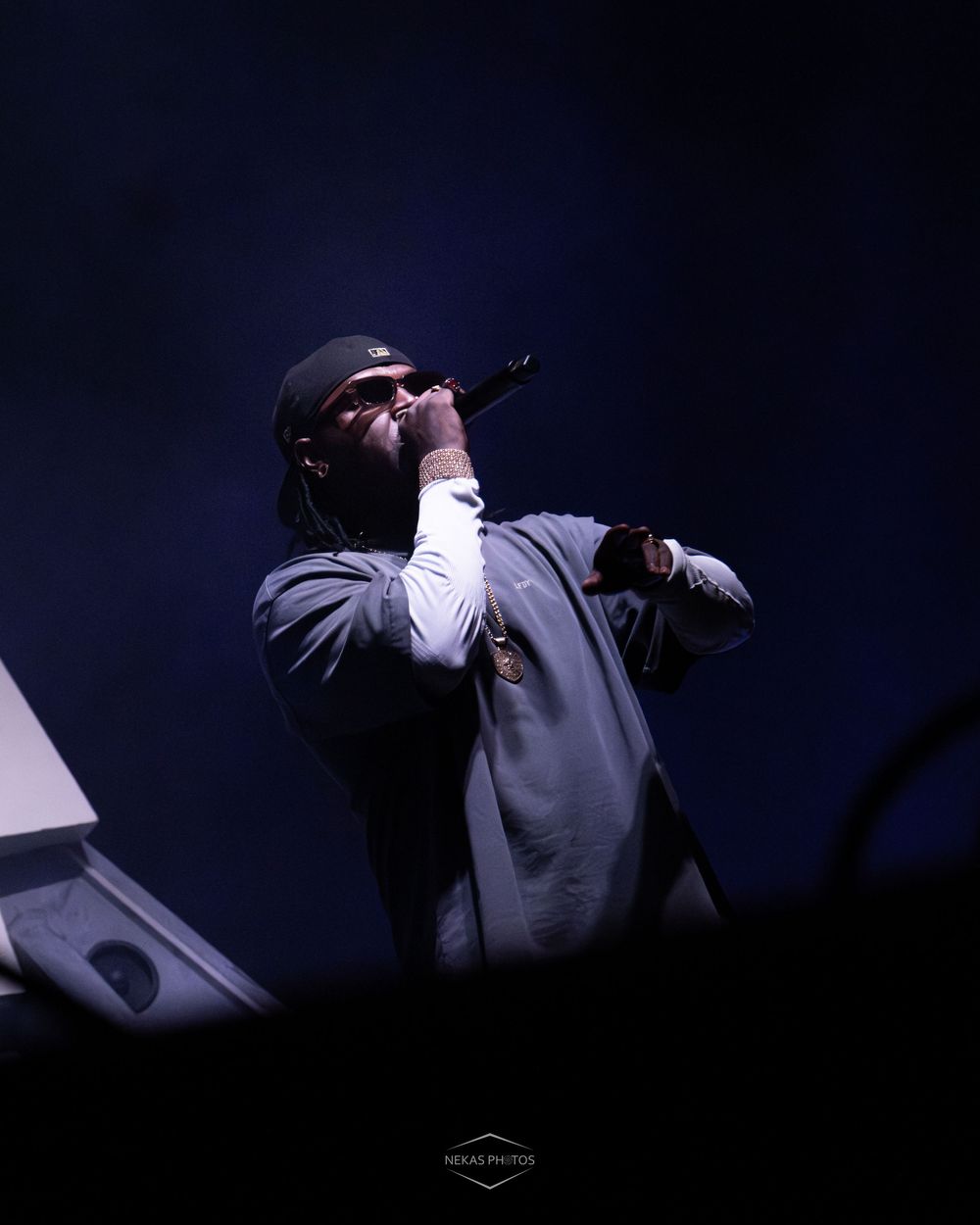 A male rapper performs on stage holding a microphone, wearing a gray jacket and black cap under dramatic blue lighting.