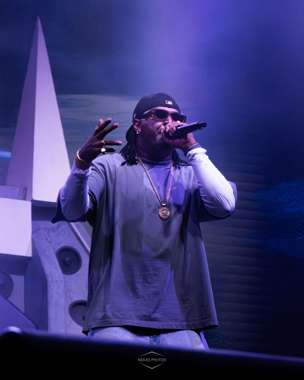 Rapper performing on stage holding microphone, wearing grey hoodie, black cap, and gold chain necklace under purple stage lights.