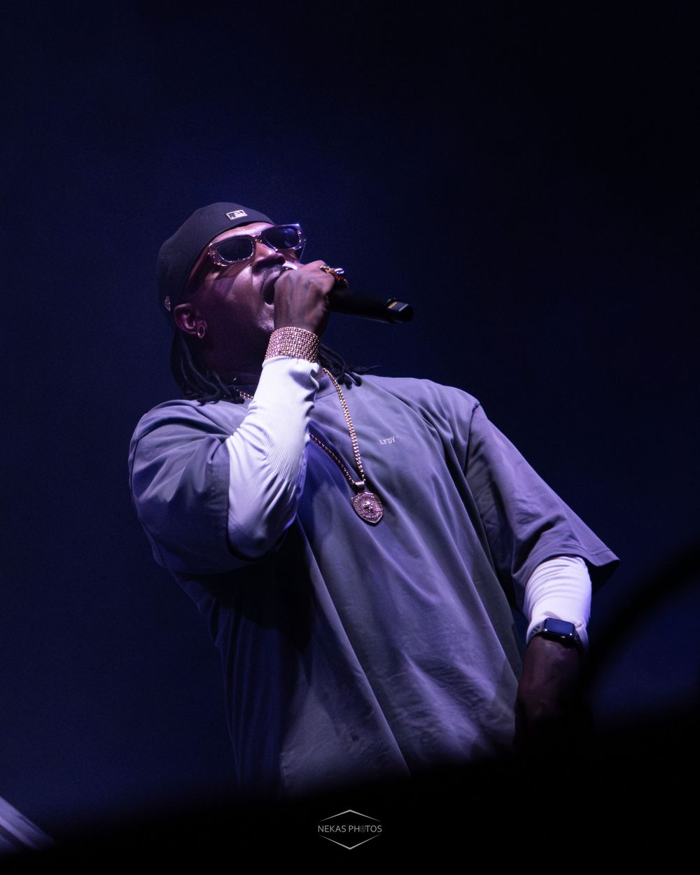 Male hip-hop artist singing into microphone on stage, wearing grey jacket, sunglasses, and gold chain under dramatic blue lighting.
