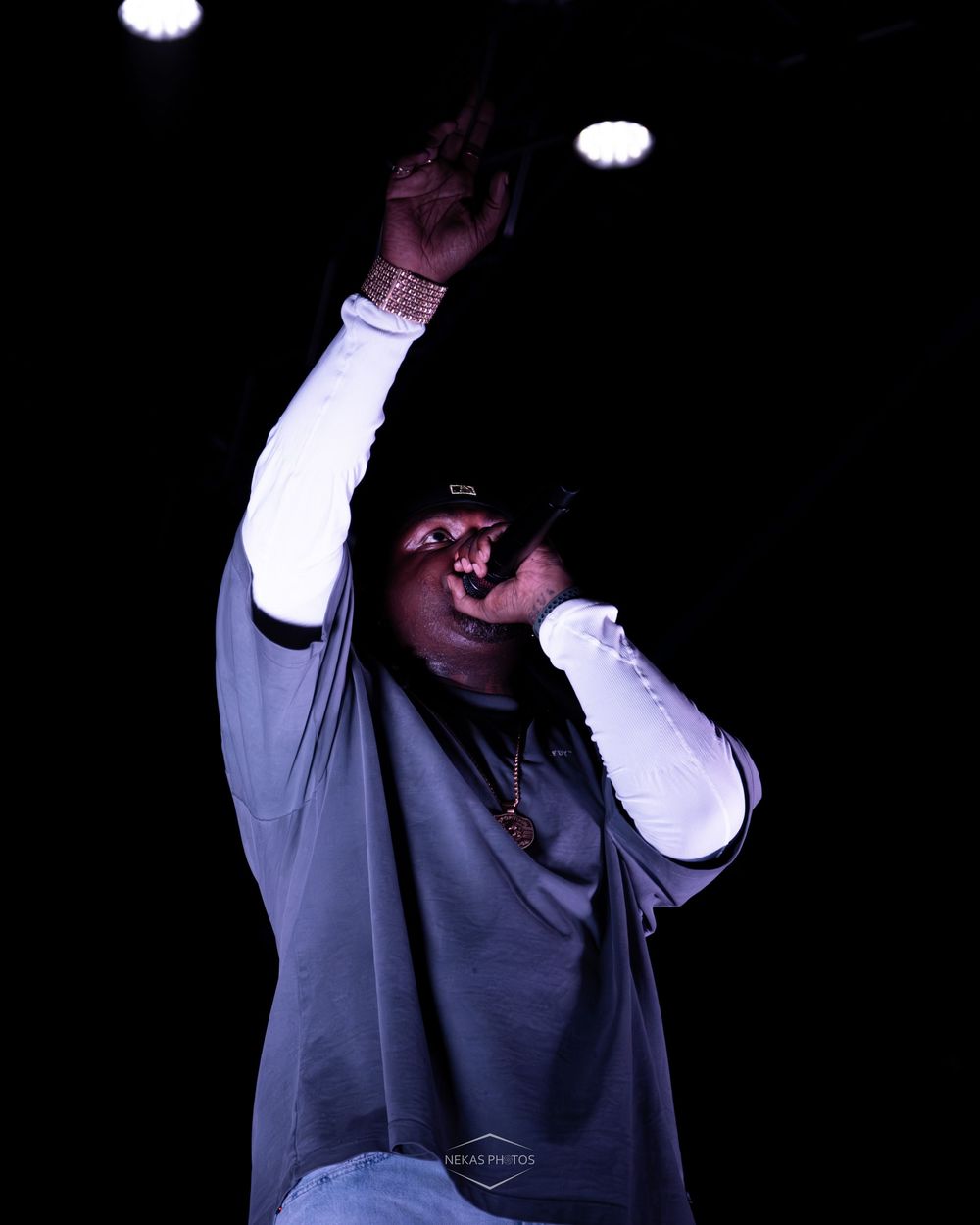 A performer on stage raises one arm dramatically while wearing a gray hoodie, cap, and gold chain under dark stage lighting.