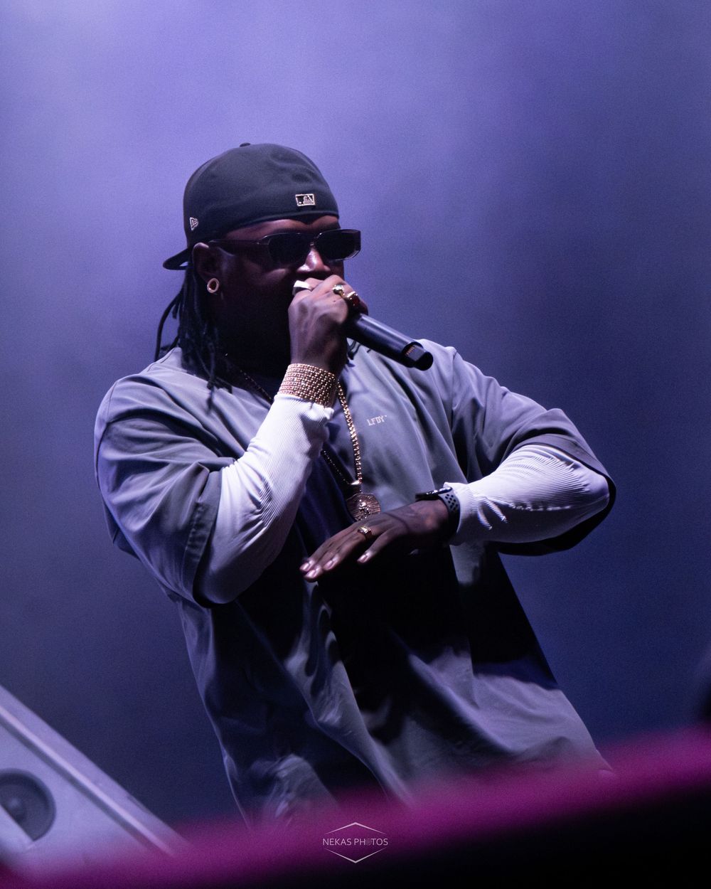 A rapper performs on stage holding a microphone, wearing a cap, sunglasses, and grey jacket under dramatic purple stage lighting.