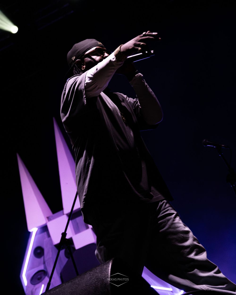 A rapper performs on stage under purple lights, holding a microphone while wearing all black attire.