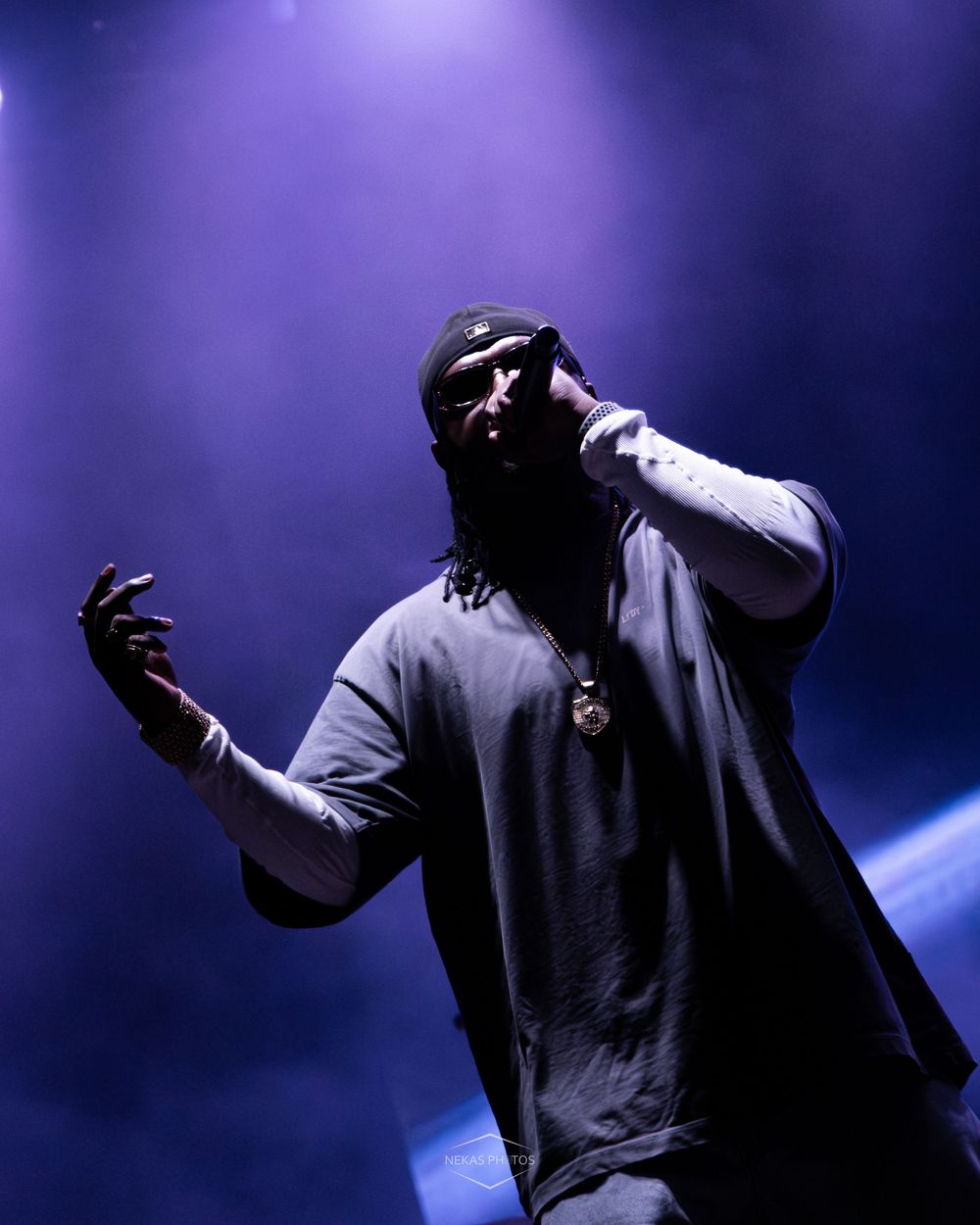 A performer on stage under dramatic purple lighting, wearing a gray oversized tee and gold chain, holding a microphone.