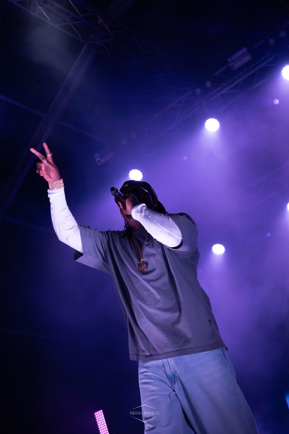 Rapper performing on stage with purple lights, holding microphone and making peace sign gesture.