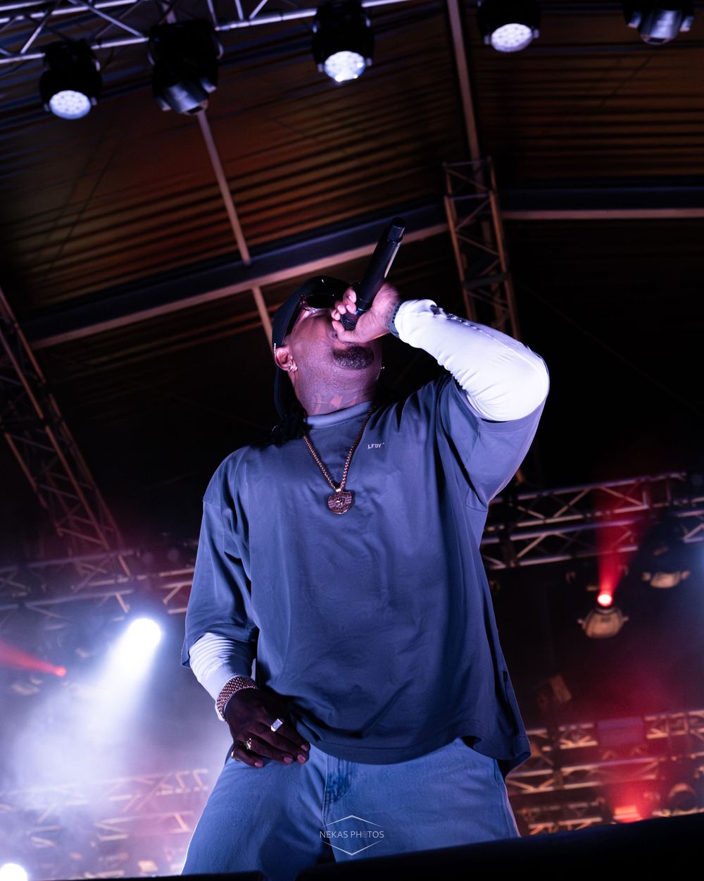 A rapper performing on stage, holding a microphone up, wearing a blue sweatsuit and gold chain under dramatic concert lighting.