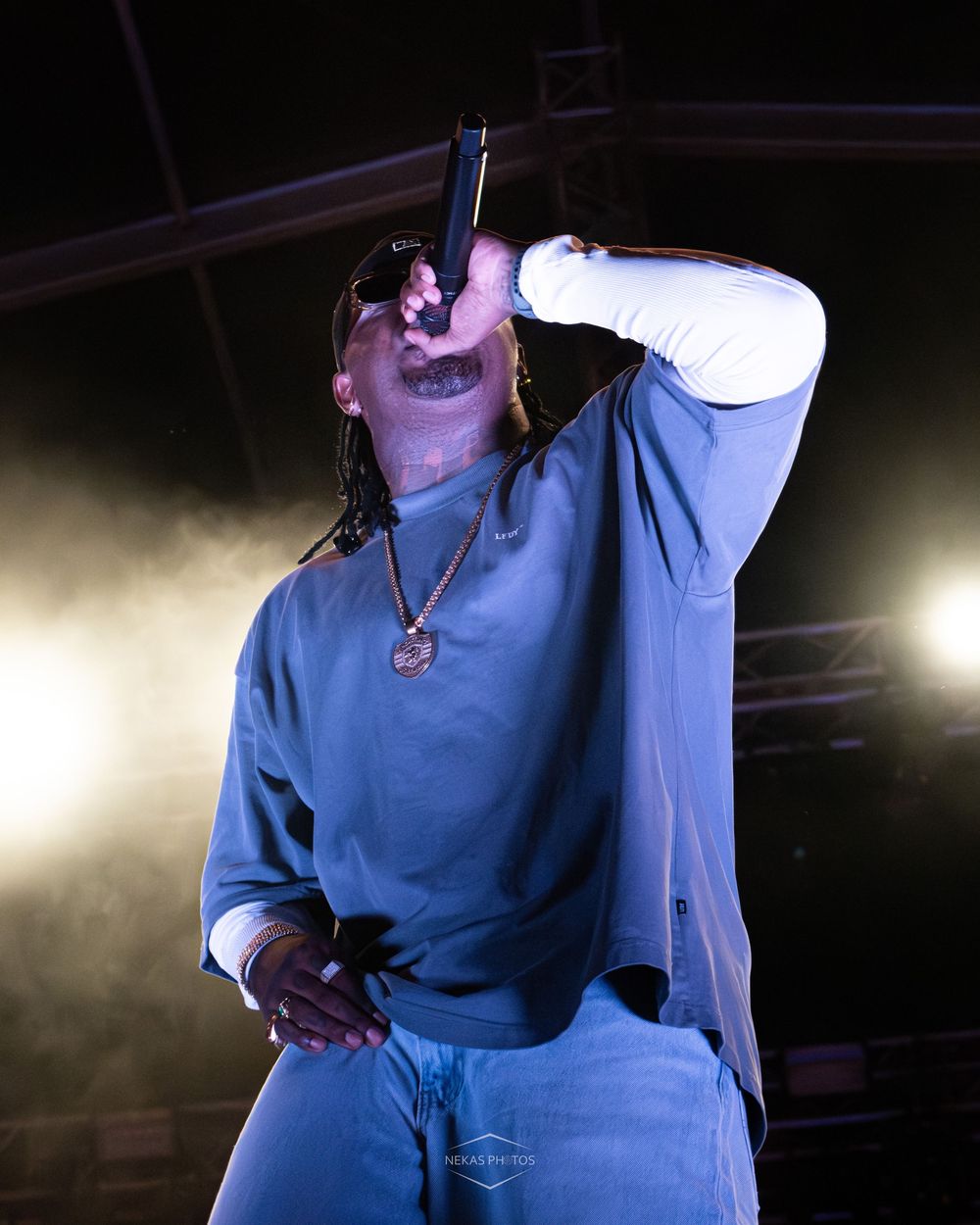 A rapper performing on stage under yellow spotlights, wearing a blue sweatsuit and gold chain, holding a microphone up while singing.