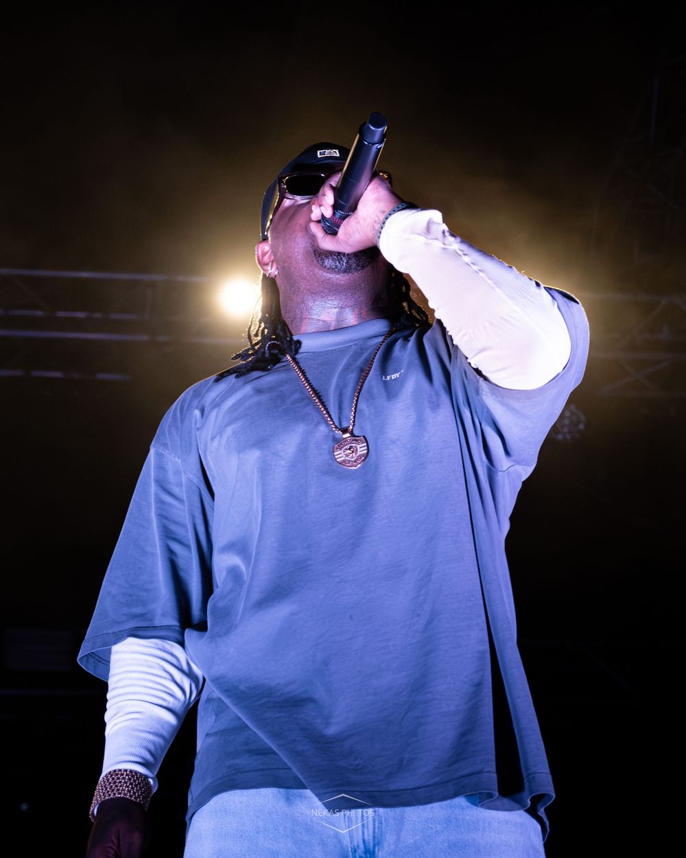 A rapper performing on stage holding a microphone, wearing a blue oversized tee, chain necklace, and sunglasses under dramatic lighting.