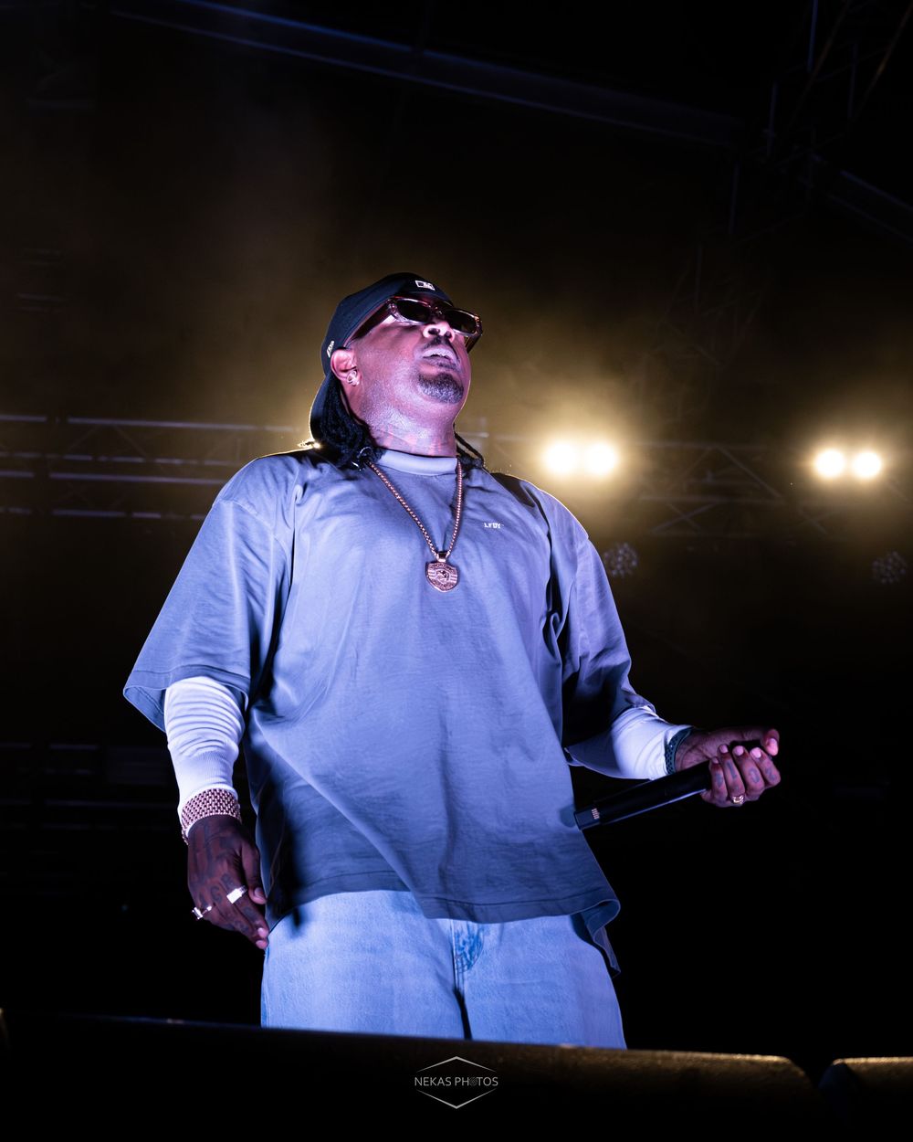 Rapper performing on stage wearing blue outfit, cap, sunglasses, and gold chain necklace under bright concert lights.