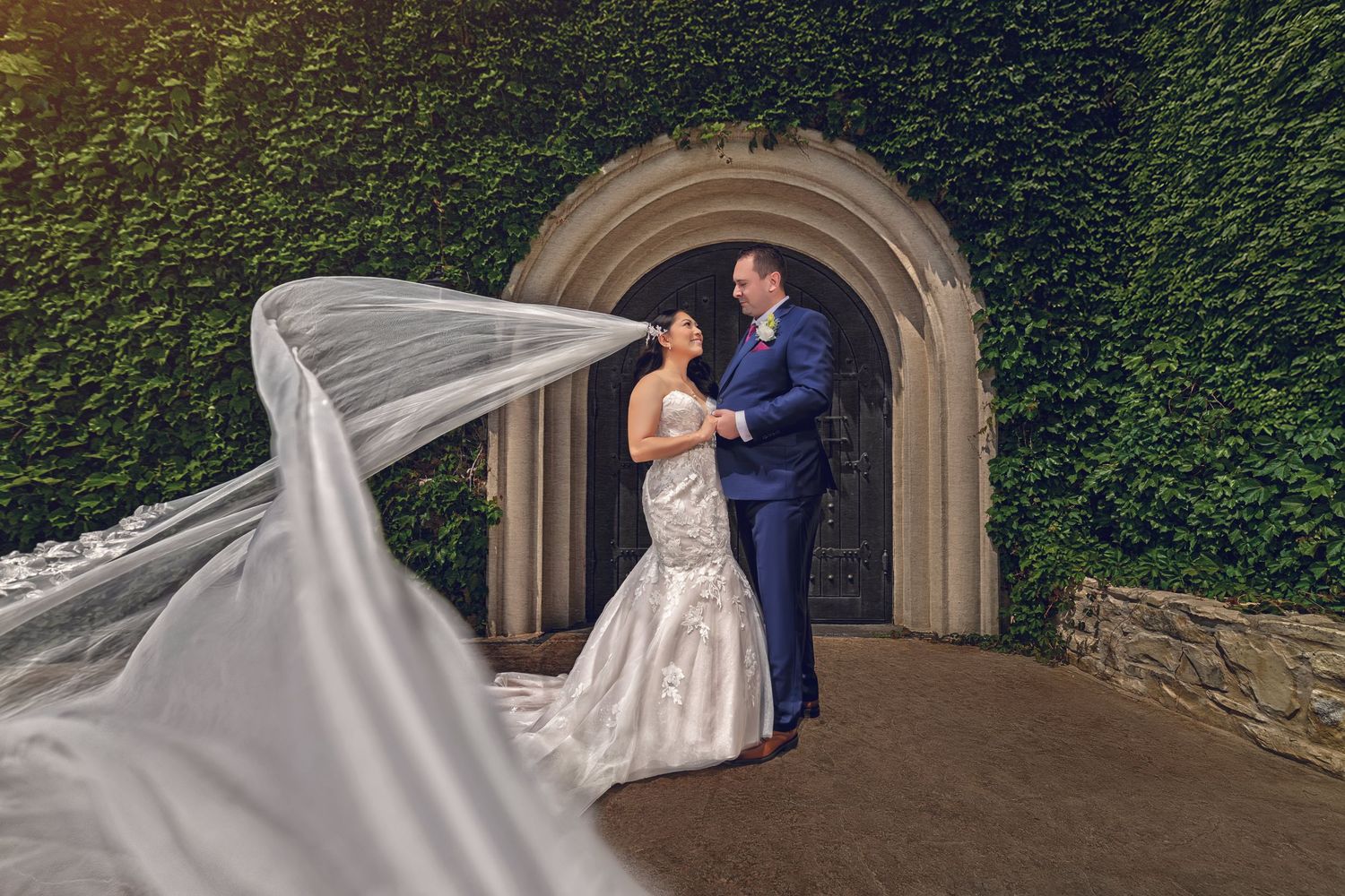 Romantic garden wedding photography in Carrollton, TX. Couple in front of an ivy-covered stone wall with a dramatic veil toss effect.