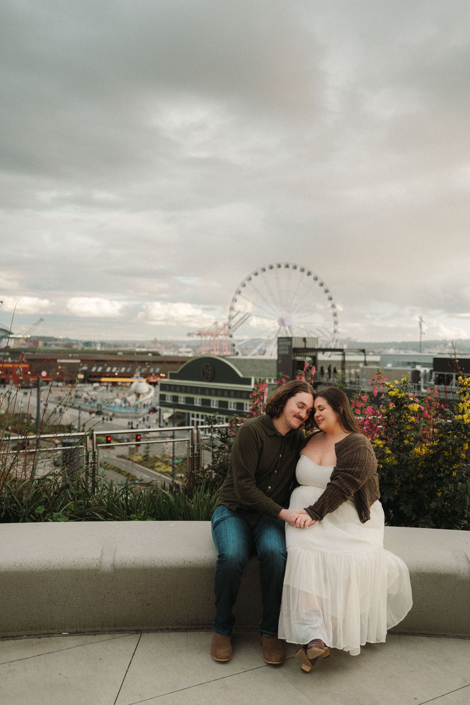 Couple sharing a tender moment on a bench with Seattle's Great Wheel and waterfront in the background.