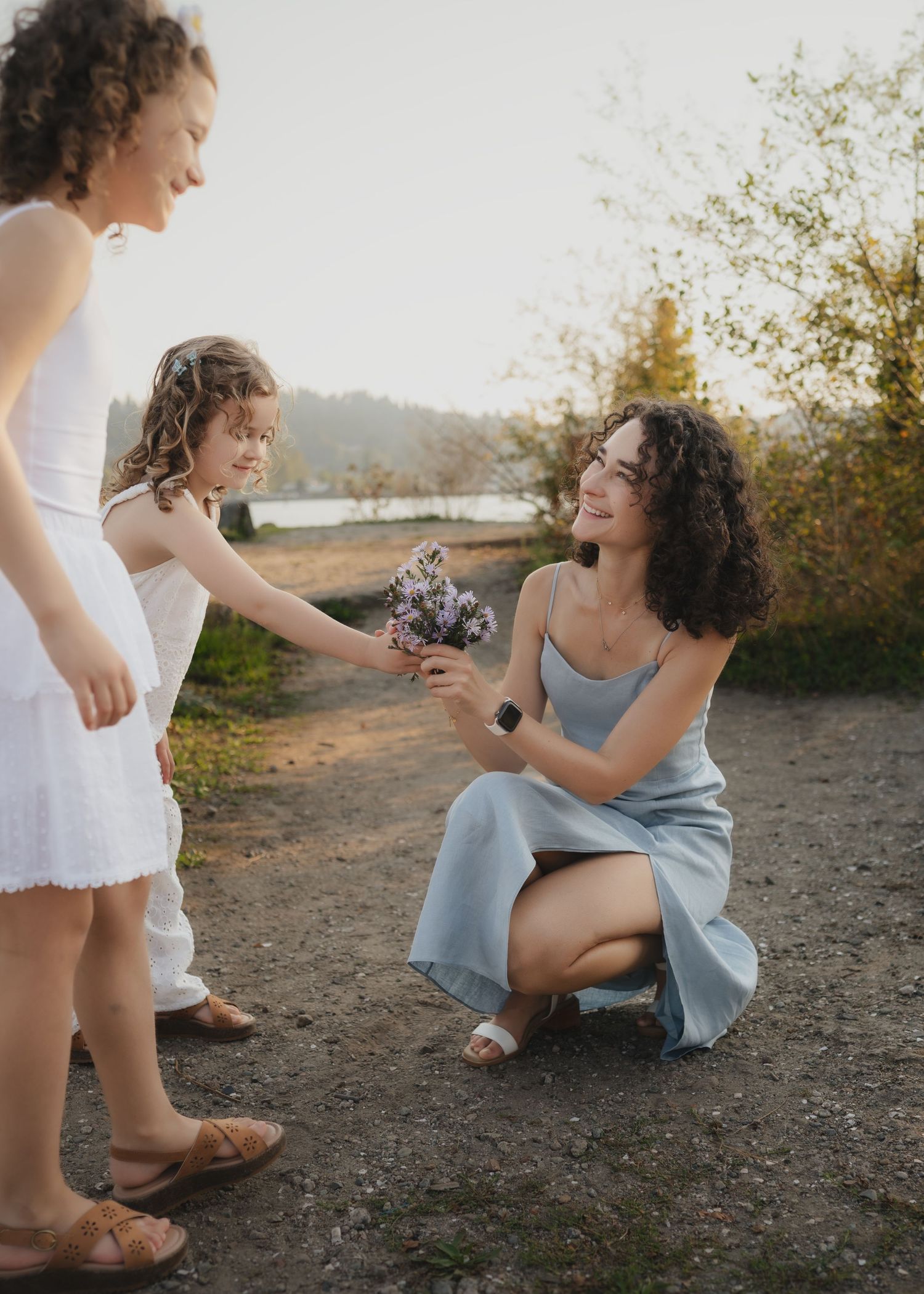 A woman crouches to receive a small bouquet of flowers from a young girl outdoors near a lake at sunset.