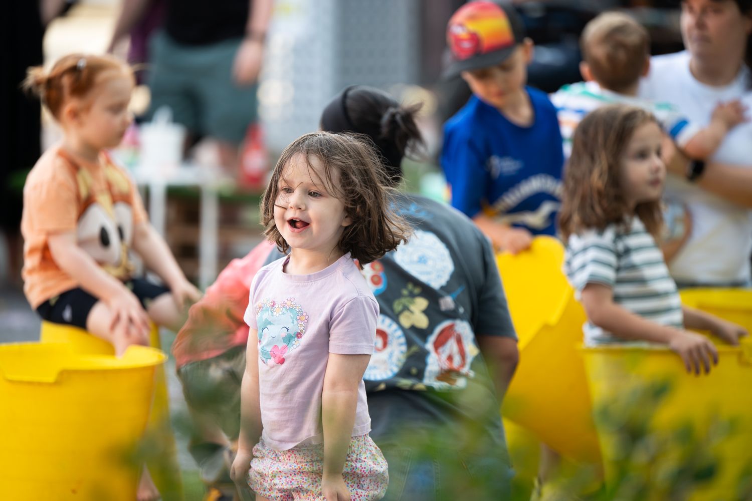 Happy children play with yellow buckets at a lively outdoor community event on a warm sunny day.