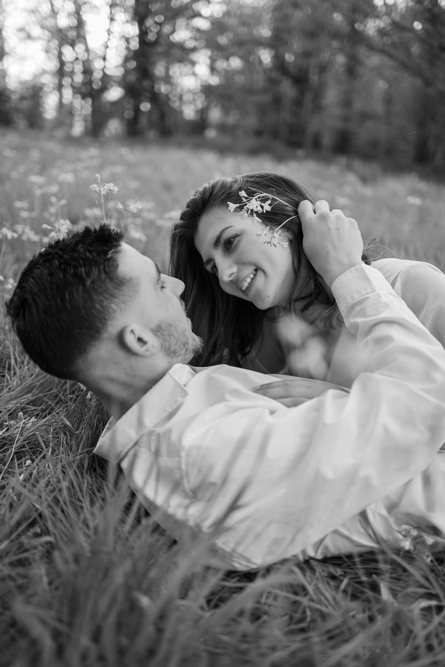 Romantic couple in white lying in green meadow with wildflowers, sharing a tender moment face to face.