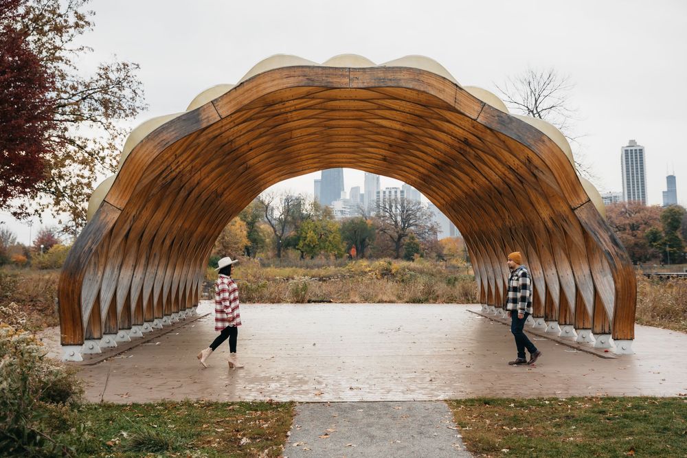 Two people walk through a wooden arched pavilion in an autumn Lincoln park with Chicago skyline in the background.