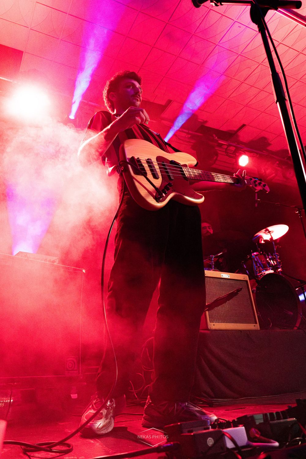Guitarist performing on a foggy stage bathed in red light with a drum kit visible in the background.